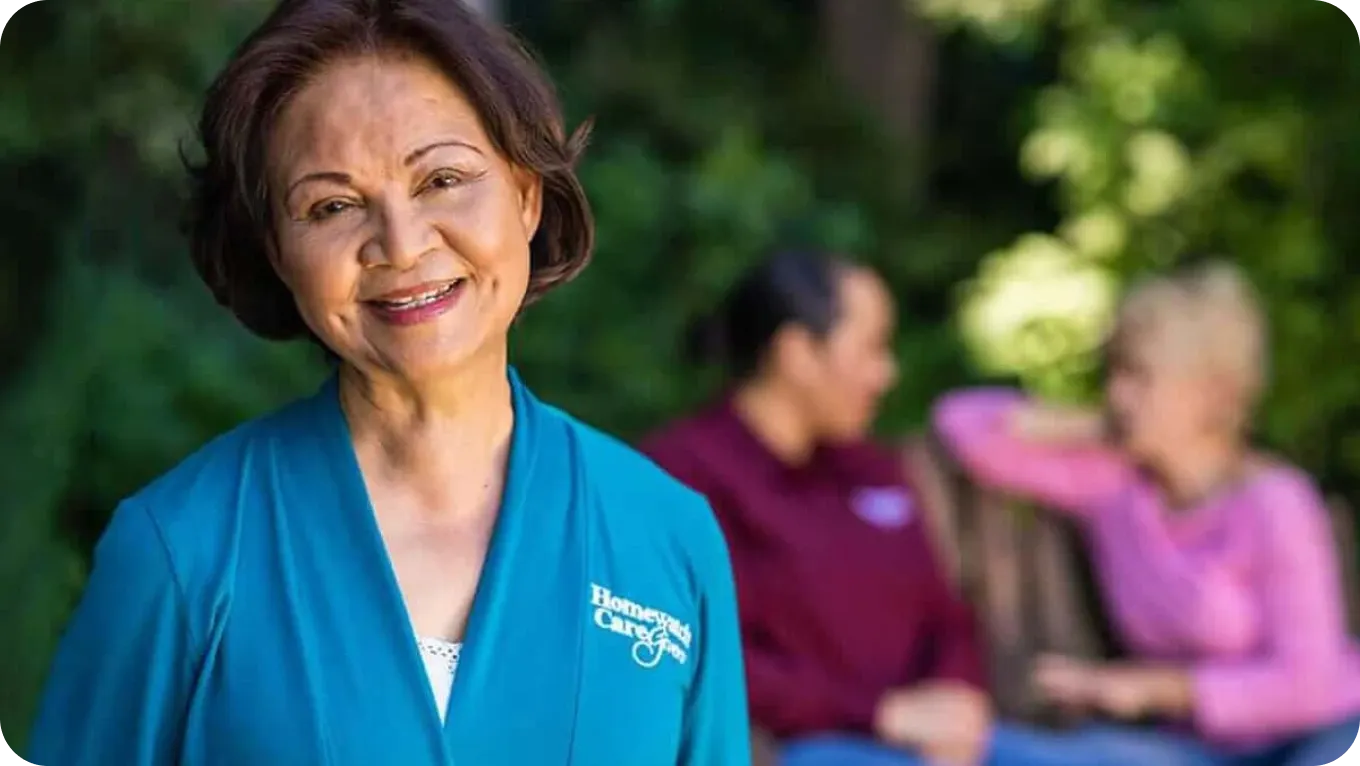 Smiling caregiver in blue Homewatch CareGivers uniform, with two women talking on a bench in the blurred background outdoors.