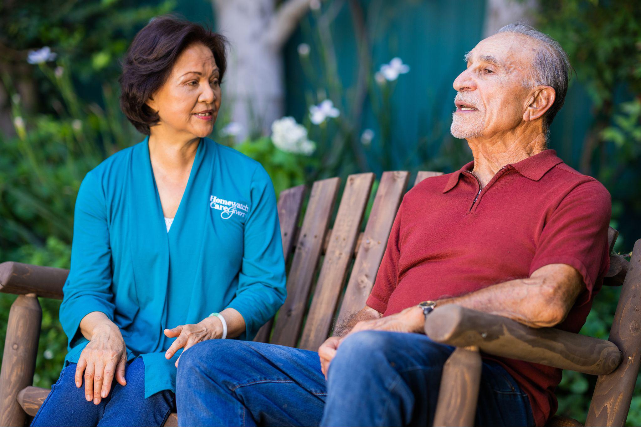 A female caregiver sitting on wood bench with senior man outside.