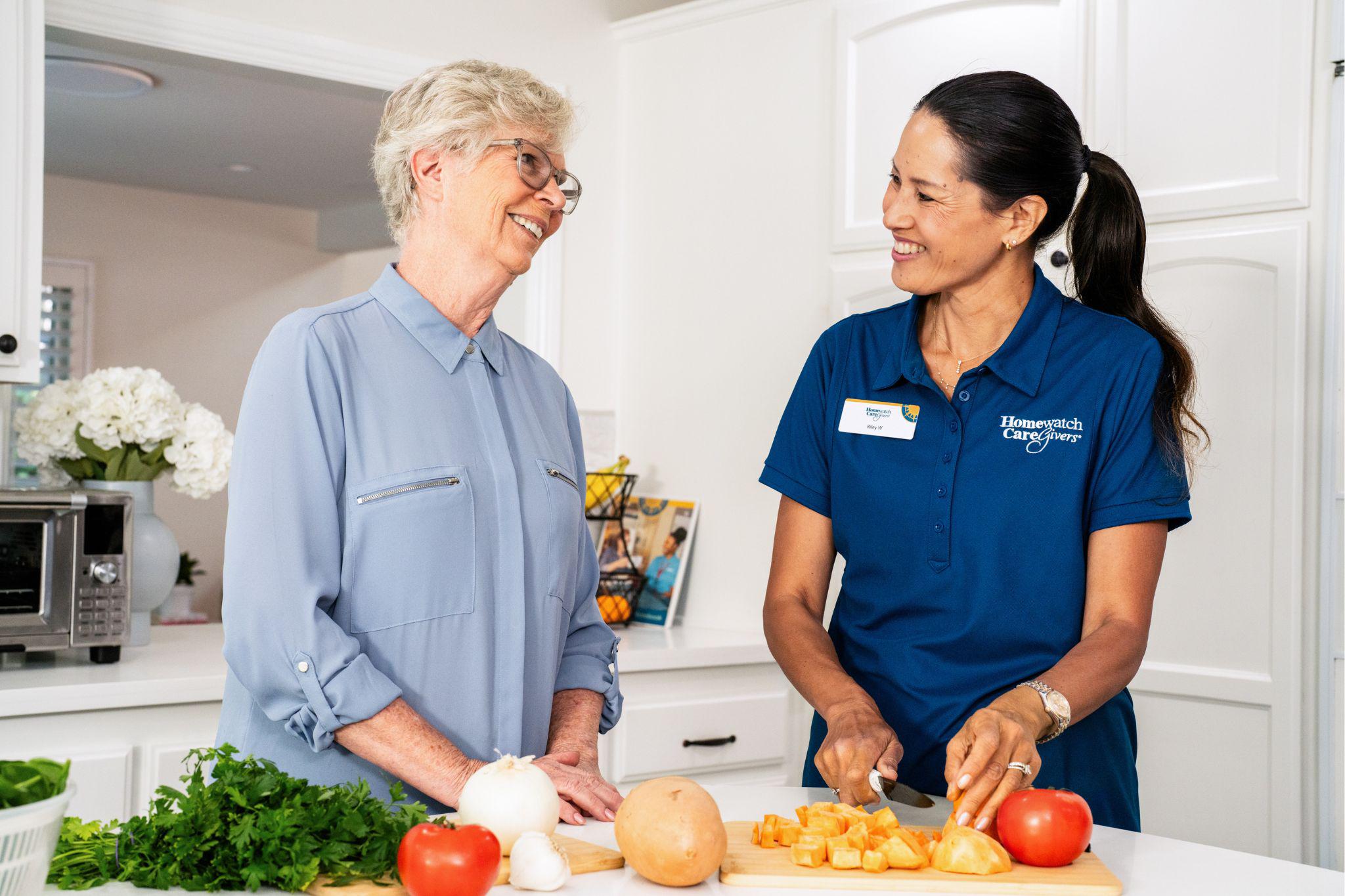 older woman standing up in her kitchen with female caregiver chopping up vegetables at kitchen island