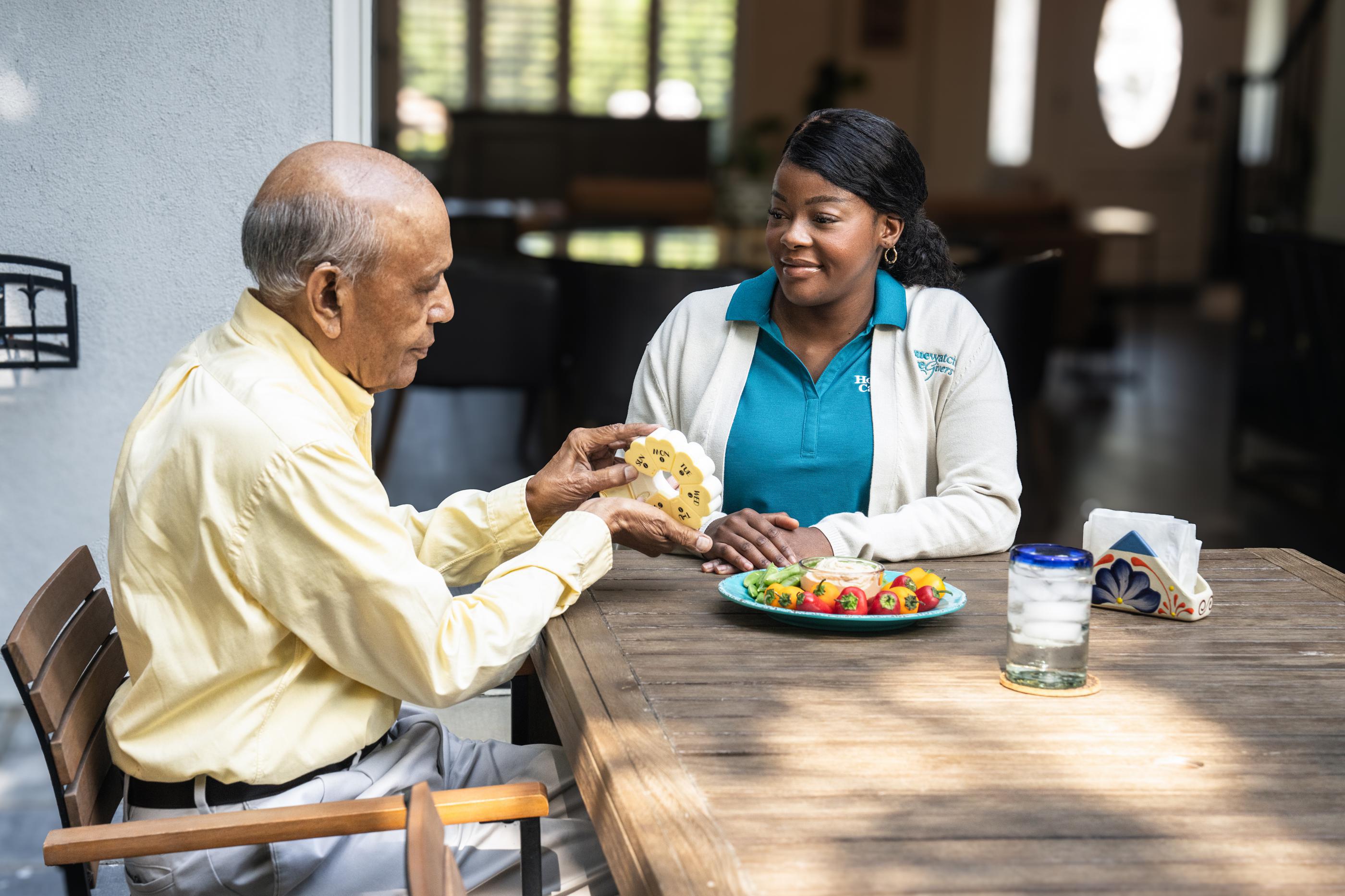 man sitting on chair at table with pill case talking to caregiver