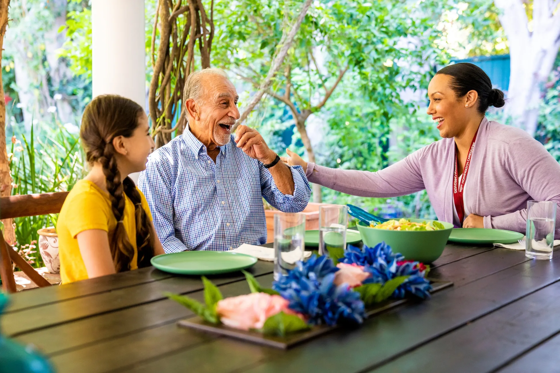 Senior man laughs, eating outdoors with his granddaughter and Caregiver at a table set for a meal.