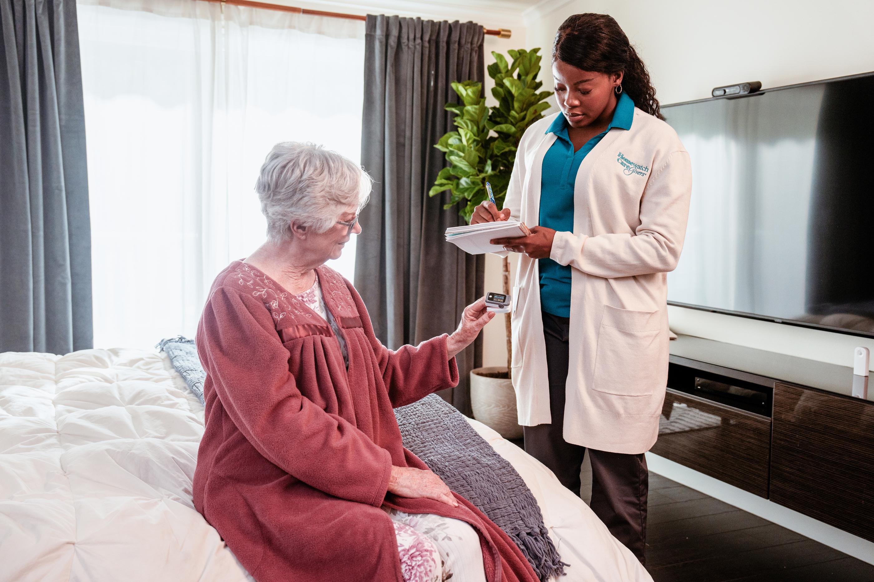 female caregiver taking blood oxygen level of elderly woman on edge of bed in her home