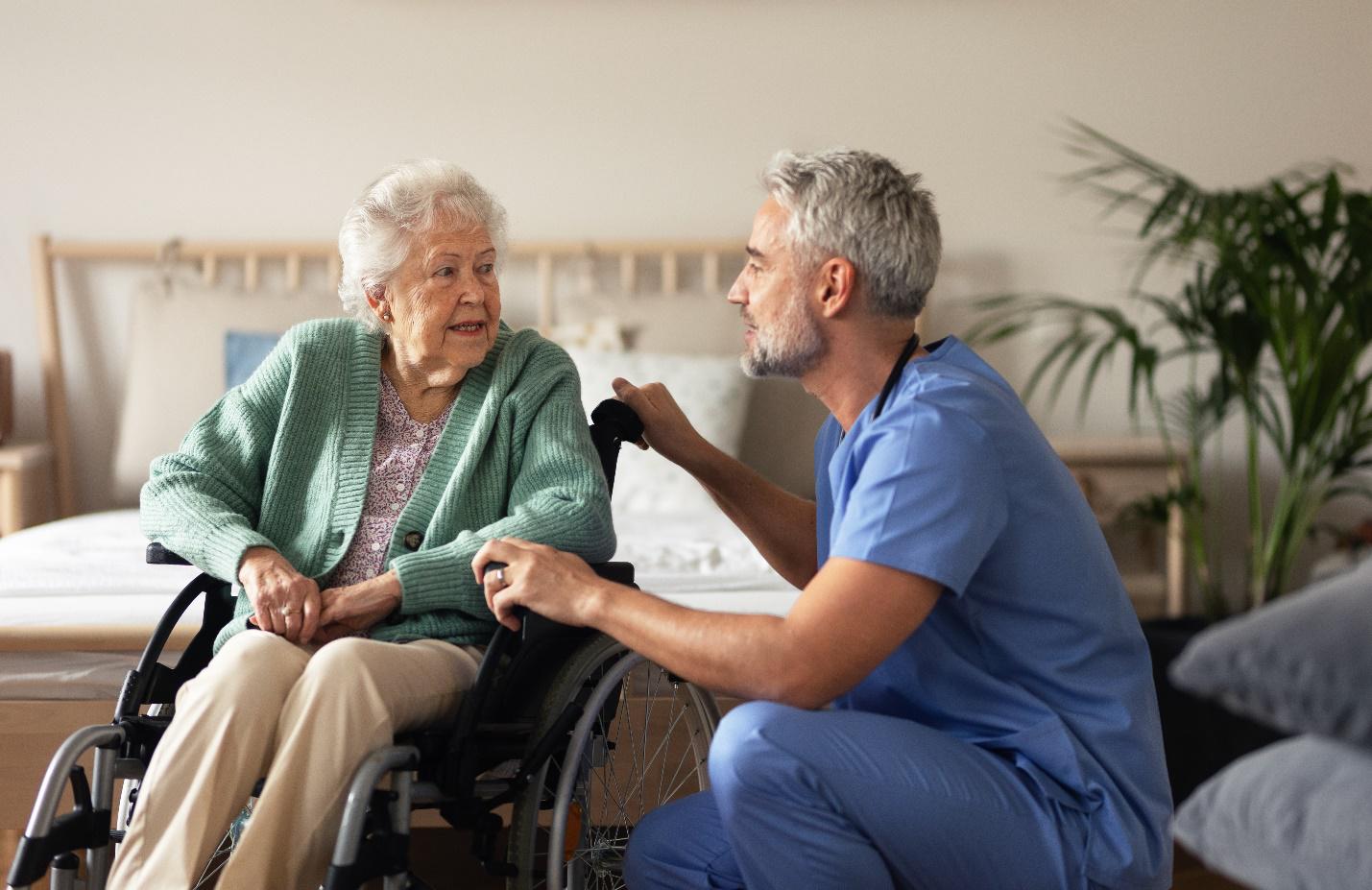 A person in a blue scrubs talking to an old person in a wheelchair
Description automatically generated